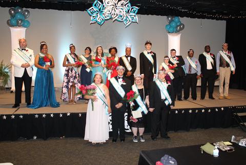 Image description: The 2019 Royal Court, a group of men and women dressed in pageant dresses and tuxes, standing on a stage that is decorated with blue and silver balloons and stars.